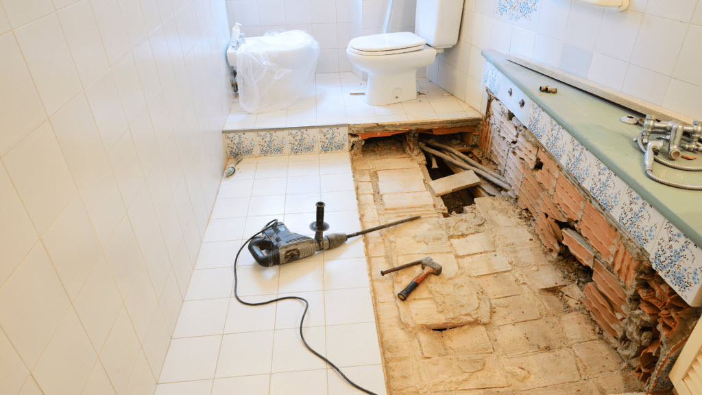 Bathroom mid-renovation with floor tiles removed, exposed pipework, a drill and hammer on site