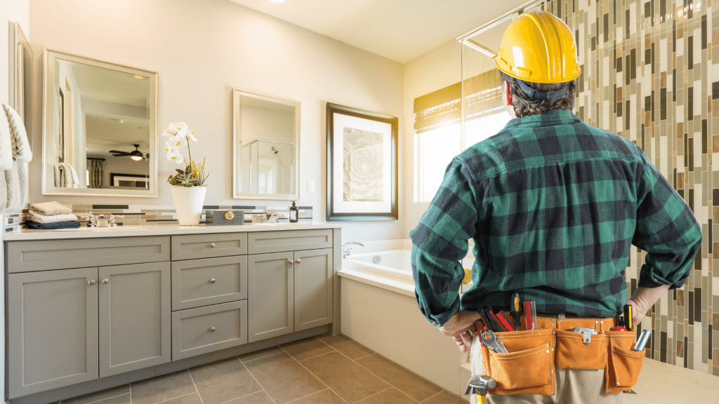 Tradesperson in a hard hat inspecting a finished bathroom with a grey vanity and mirrors