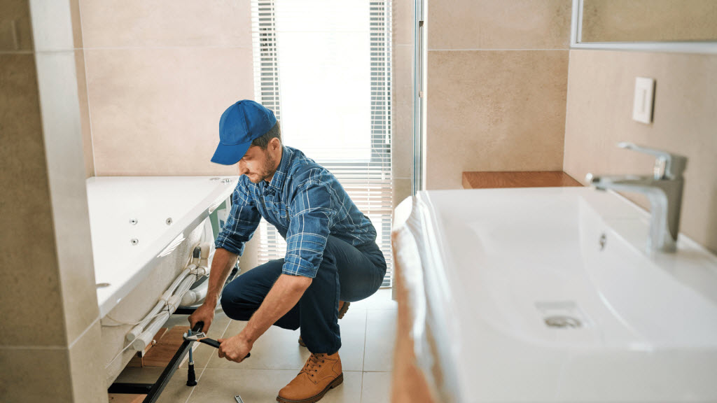 Plumber in a blue cap kneeling to fit pipework beneath a bathtub next to a sink vanity