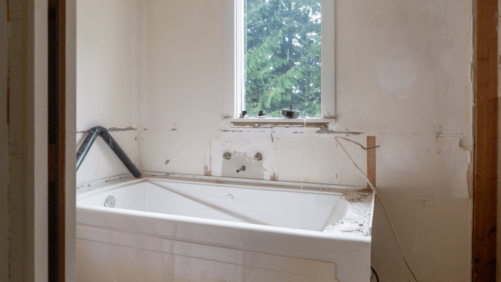 Bathtub installed in a partly finished bathroom with bare walls, a window, and exposed plumbing