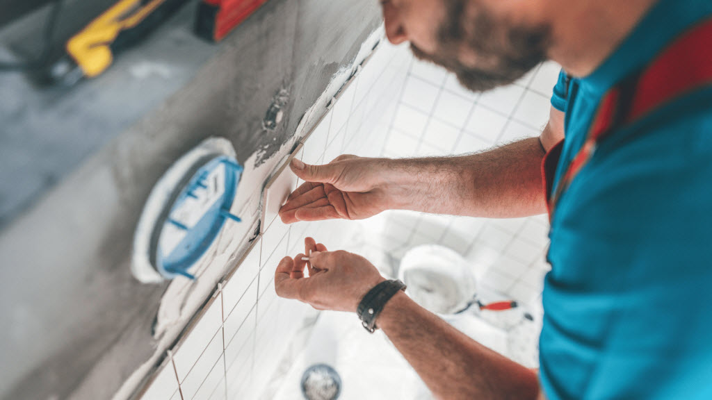 Person applying a mosaic wall tile above a chrome tap and sink during a bathroom retiling job