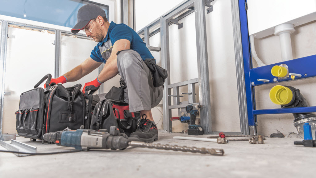 Tradesperson kneeling to pack tool bags beside exposed stud framing and plumbing pipework on site
