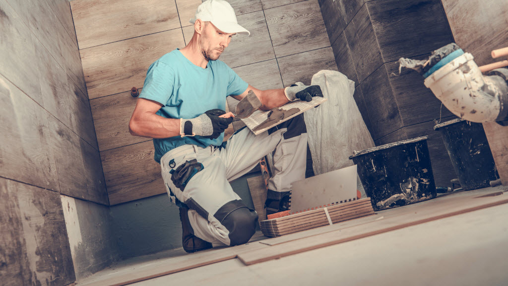 Tiler kneeling to fit tiles to a bathroom wall, surrounded by grout tubs and cut tile pieces