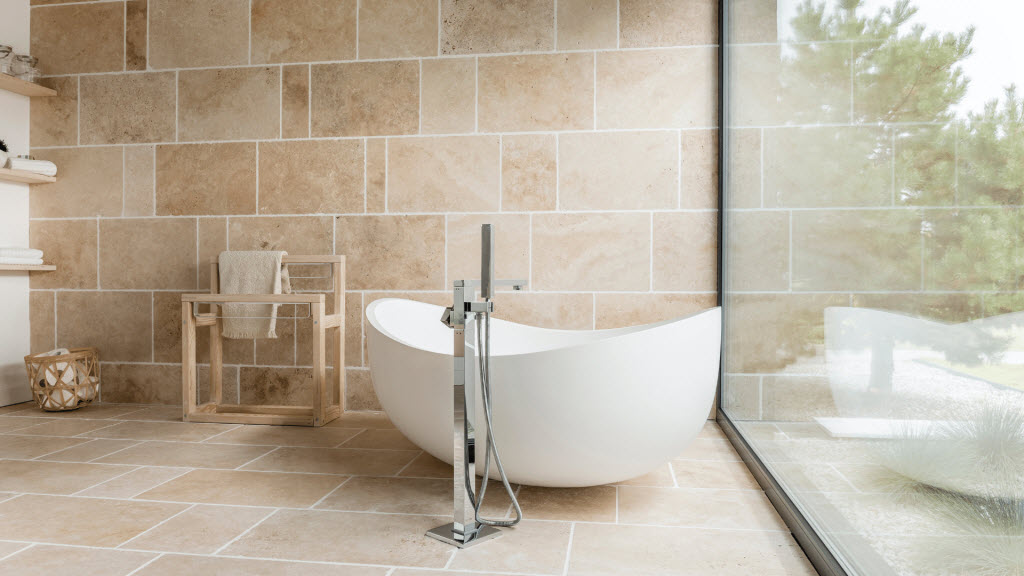 Freestanding white bathtub with a floor-mounted tap against a stone-tiled wall and window